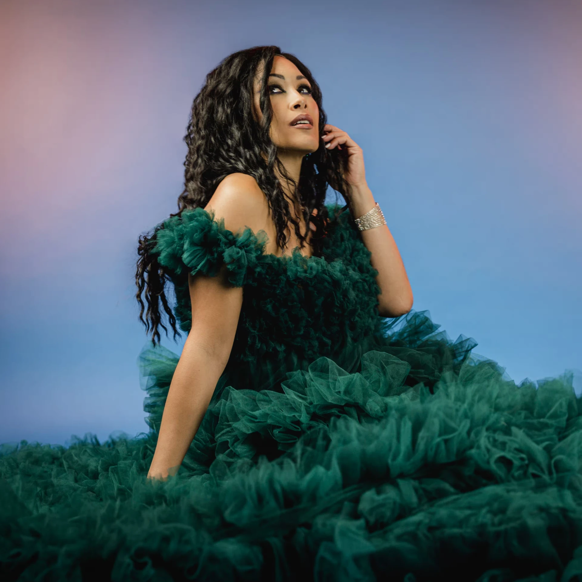 Melina Soochan seated in a sculptural emerald gown against a blue backdrop